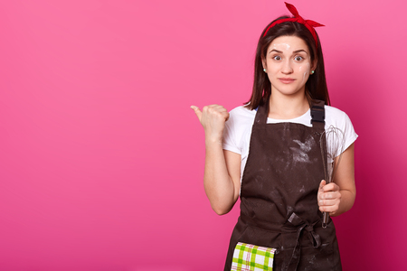 Charismatic beautiful young housewife posing isolated over pink background in studio, looking directly at camera, showing direction with her thumb, looks impressed.の写真素材