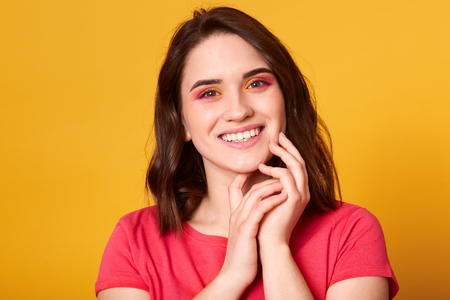 Indoor shot of beautiful female with natural perfect skin and bright make up in pink and orange colours, looking smiling at camera, keeps hands together near face, dressed red t shirt. Beauty concept.の写真素材