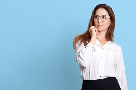 Portrait of young pensive woman touching her chin, has something in mind, posing with thoughtful facial expressions, wearing white shirt and black skirt, isolated over blue background. Copy space.の写真素材