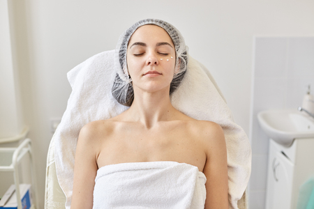 Close up portrait of well kept beautiful woman laying on coach with closed eyes, having peaceful facial expression, being naked, rolled with white towel and medical cap, cream points under eyes.の写真素材