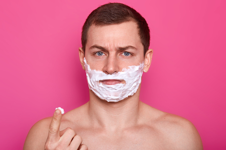 Close up portrait of confused handsome young man with foam on his face looking directly at camera while standing against rose background, points forefinger with shaving cream up. Skin care concept.の写真素材