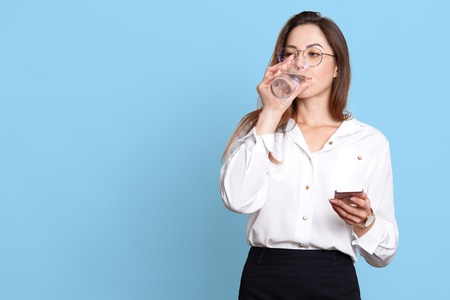 Beautiful young woman with healthy skin holding mobile phone in hand and drinking clean water while having business conversation, business lady wearing white blouse and black skirt. Copy space.の写真素材