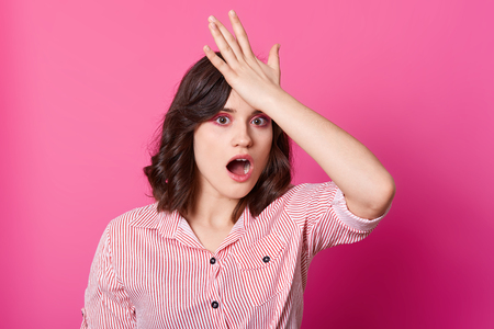 Studio shot of worried forgetful beautiful brunette woman in stylish striped shirt, looking at camera with opened mouth, holding palm on forehead, being shocked, standing isolated over rosy backgroundの写真素材