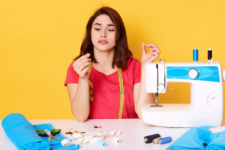 Indoor shot of upset busy young woman in red t shirt sitting at desk full of sewing tools and holding torn thread in workshop, model posing isolated on yellow. Fashion, sewing and hobby concept.の写真素材