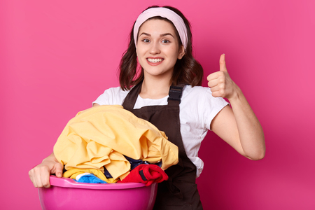 Close up portrait of good looking slender housewife holding pink basin with clean clothes, having positive facial expression, showing super sign with her fist and thumb, looks emotional and happy.の写真素材