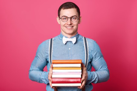 Smiling positive man holds bunch of colorful books in both hands, posing isolated over pink background in studio. Athletic strong man wears blue shirt, stylish spectacles, bow tie and suspenders.の写真素材