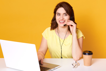 Half lengh photo of woman operator sitting at office desk, looking aside, touching headset, isolated over yellow background, brunette female wears casually, looks cheerful. Call center concept.の写真素材