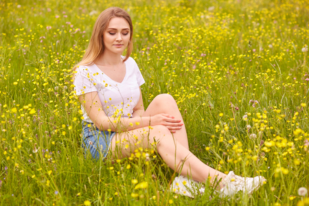 Close up portrait of young model blonde woman sitting on grass, posing in white casual t shirt and denim skirt, has romantic expression, siting on ground surrounded field flowers and looking down.の写真素材