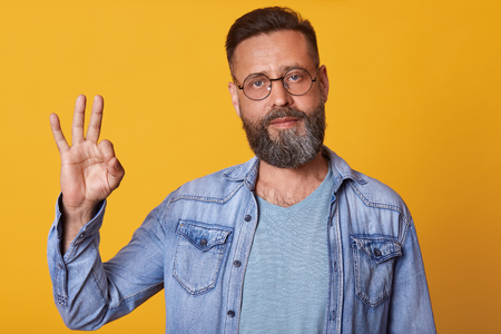 Handsome man with beard wearing denim jacket and gray t shirt stands against yellow studio wall and showing ok sign with hand and fingers, has satisfied facial expression. Body language concept.の写真素材