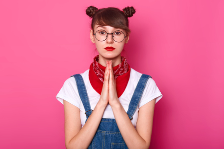 Portrait of black haired brown eyed young female standing isolated over pink background, praying with putting hands together, looking directly at camera, having bunches and bright colored lips.の写真素材