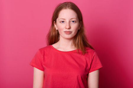Half length portrait of young student female looks with calm facial expression at camera, wearing red casual t shirt, has long straight brown hair and beautiful blue eyes, isolated over pink backgroundの写真素材