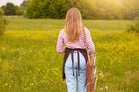 Back view of female artist finishing her picture on sketchbook, standing in front of beautiful nature, wearing striped shirt, pants and brown apron, female has long blonde hair. Art and creating concept.の写真素材
