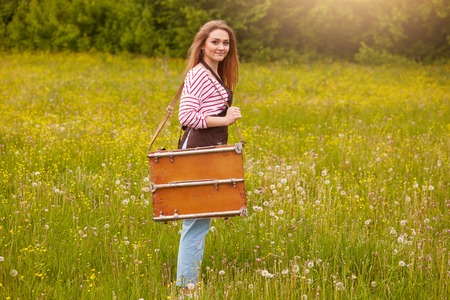 Outdoor shot of young artist standing with drawing easel in open air, full length photo of beautiful girl in striped shirt and apron ready to paint landscape, female with fair hair looks at camera.の写真素材