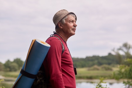 Close up profile shot of elderly man with backpack, wearing red casual sweater and cap, standing near river and looking far away, senior male likes to spend time in active way. Backpacking concept.の写真素材