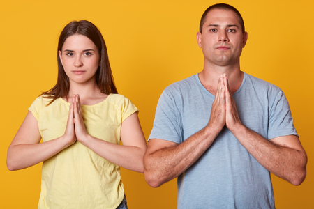 Studio shot of grateful thankful young couple holding hands in praying gesture, sincere love, man and woman directly at camera, have serious facial expressions, isolated over yellow studio background.の写真素材