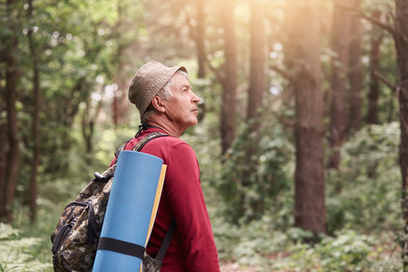 Profile of senior man looking aside attentively, going camping, wandering in forest, uniting with nature, having backpack and sleeping pad at his back, spending time at fresh air. Wanderlust concept.の写真素材