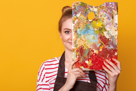 Indoor studio shot of satisfied talented girl holding palette to mix colors, covering half of her face with it, looking directly at camera, ready to paint masterpiece, being inspired by her hobby.の写真素材