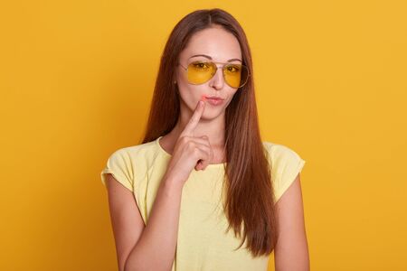 Close up portrait of beautiful dark haired woman points to her lips with fore finger, has virus, being infected with col sore herpes, needs treatment, dressed casually, isolated over yellow backgroundの写真素材
