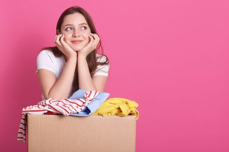 Horizontal shot of cheerful young volunteer standing isolated over rosy studio background, posing near carton box full of donated clothes. Smiling woman looking aside, keepa hands under chin.の写真素材