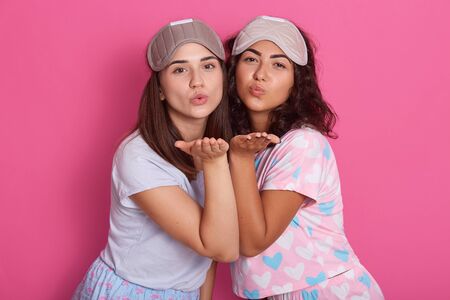 Photo of beautiful girls posing in pajamas. Studio shot of two friends standing on pink background and shows air kisses, having sleep masks on heads, likes spending time together. Friendship concept.の写真素材