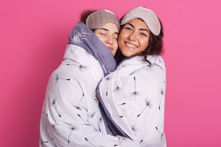 Indoor shot of smiling women with sleep masks, wearing blanket stands hagging isolated over rose studio background, having pajamas party, hagging each other, have good mood, feel happy. Friendshipの写真素材