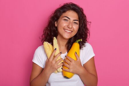 Picture of cheerful attractive model looking directly at camera, holding corn cobs and squash, keeping close to body, having black curly hair, posing isolated over pink background in studio.の写真素材