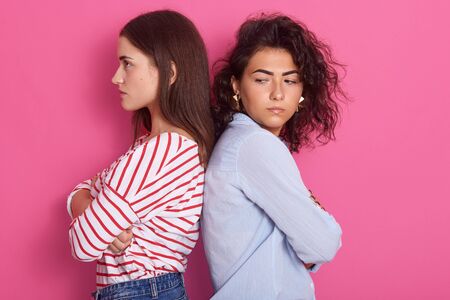 Profile portrait of beautiful girls with dark hair frowning her face in displeasure, wearing casual shirts, keeping arms folded, do not talk to each other, posing isolated over pink studio background.の写真素材