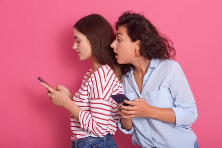 Horizontal shot of two women sharing social media videos in smart phone indoors, attractive dark haired female looks at friend's phone screen with interest and widely opened mouth. Gossip concept.の写真素材