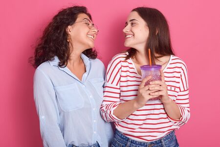 Indoor shot of two attractive adult girls standing in studio, wearing casual clothing, looking smiling at each other, posing isolated over pink background, females expressing happyness and joy.の写真素材
