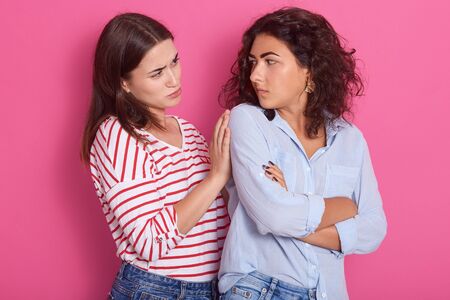 Horizontal shot of young Caucasian female with dark hair puffing cheeks, pouting, having annoyed look, feeling displeased, looks at her girlfriend, another girl touches her shulder. Same sex love.の写真素材