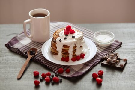Image of tasty pancakes with blueberries and raspberries on white plate, on dark brown colour rustic backdrop, cup of tea or coffe, spoon, pieces of chocholate. Delicious pancackes for breakfast.の写真素材