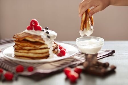 Side view of traditional family pancakes with sour cream and fresh berries isolated on dark background, woman's hand dipping muffin into sour cream, delicious dessert on checkered cotton towel.の写真素材