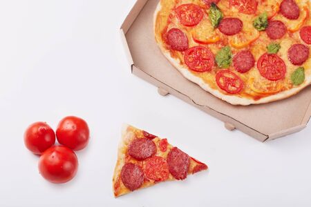 Photo of pepperoni pizza with mozzarella cheese, salami, pepper, spices and fresh spinach, cardboard box, piece of pizza and three tomatoes isolated on white background. Fast food eating concept.の写真素材