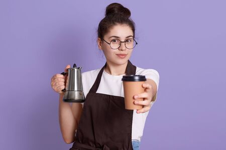 Brunette lady with hair bun wearing eyeglasses white t shirt and brown apron, holding coffee pot and offers hot drink intake away cup, standing against lilac wall.の写真素材