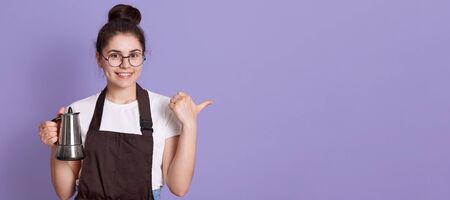 Woman with charming smile wearing eyeglasses, casual t shirt and brown apron, pointing aside with thumb, looks at camera. Copy space for advertisement and promotional text.の写真素材
