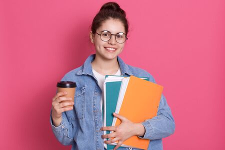 Good looking student girl posing isolated over rosy background, lady wearing denim jacket and eyewear, female holding paper folder and take away coffee, student has coffee break.の写真素材