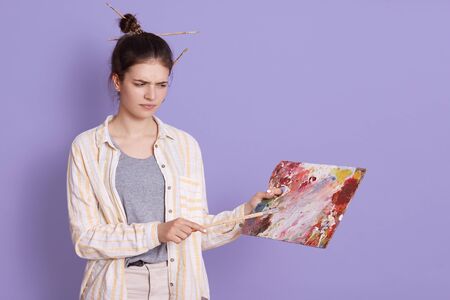 Upset artist holding picture and looking at it with upset facial expression, posing against lilac studio wall, girl wearing casual clothing, sees mistake on her work.