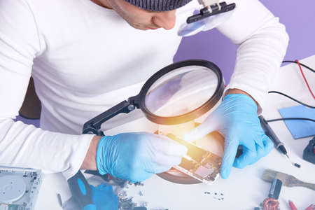Master of gadget repair service holds phone with black blanck screen for advertisement or promotion, technician wears white shirt and gray cap posing isoalted over lilac background. Technology concept.の写真素材