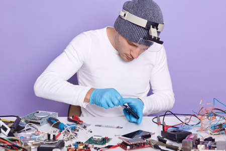 Master of gadget repair service holds phone with black blanck screen for advertisement or promotion, technician wears white shirt and gray cap posing isolated over lilac background. Technology concept.の写真素材