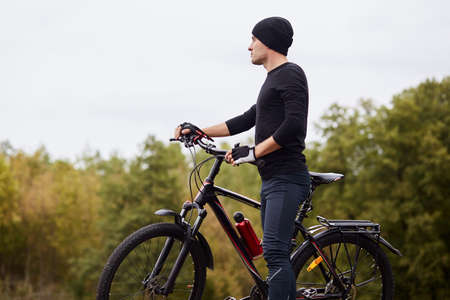 Outdoor shot of man riding bike in forest around trees, handsome man wearing black sportwear and...