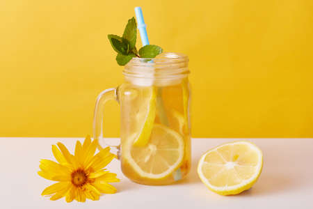 Iced tea with lemon slices and mint, glass pitcher with citrous refreshing summer cocktail and drinking straw on table decorated with flower and half of lemon.の写真素材