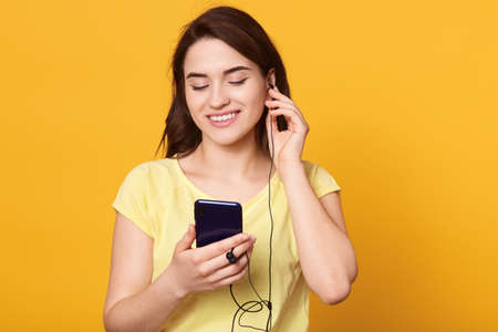 Smiling brunette female listening to music via modern smart phone and earphones, looking at mobile phone's display, lady in casual clothing posing isolated over yellow background.の写真素材