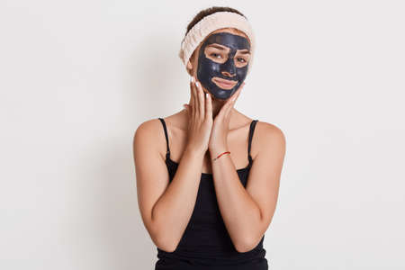 Pleased european woman applies nourishing clay mask on face, has glad expression, looks directly at...
