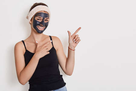 Portrait of happy cheerful woman improves her face skin, applies peeling mask, being in high spirit, models posing against white wall and pointing with both hands aside.の写真素材