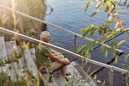 Man sitting on stairs of wooden placing waiting for fish to bite on set rods, looking at beautiful...
