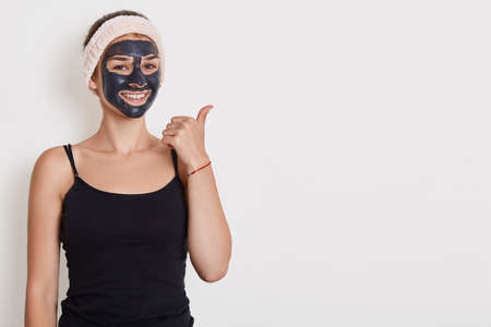 Photo of smiling woman wears black t shirt and hair band with facial mask, has beauty procedures at home, positive expression, points aside with thumb isolated over white wall.の写真素材