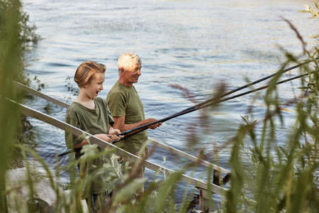 Fishing by lake, grandson and grandfather near river, holding fishing rods in hands, look excited...