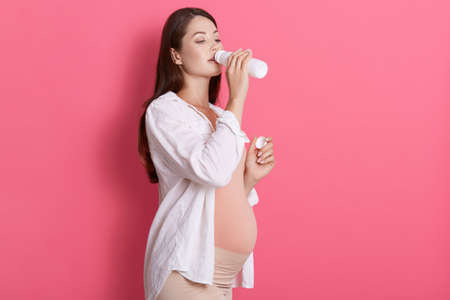 Attractive pregnant girl drinks milk from bottle isolated over pink background, lady with brown hair...