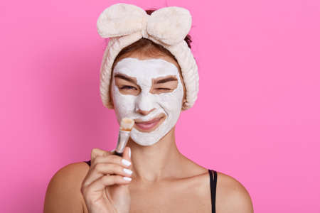 Young beautiful woman with clay facial mask on her face holding brush in hands after doing beauty treatment, making funny face while does skin care manipulation, having fun at home.の写真素材