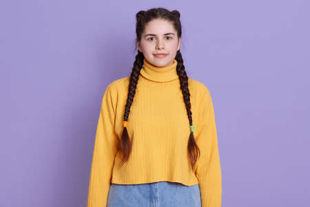 Portrait of beautiful cute girl with pigtail hairstyle, wearing yellow shirt isolated over lilac background, having calm facial expression, looks at camera.の写真素材